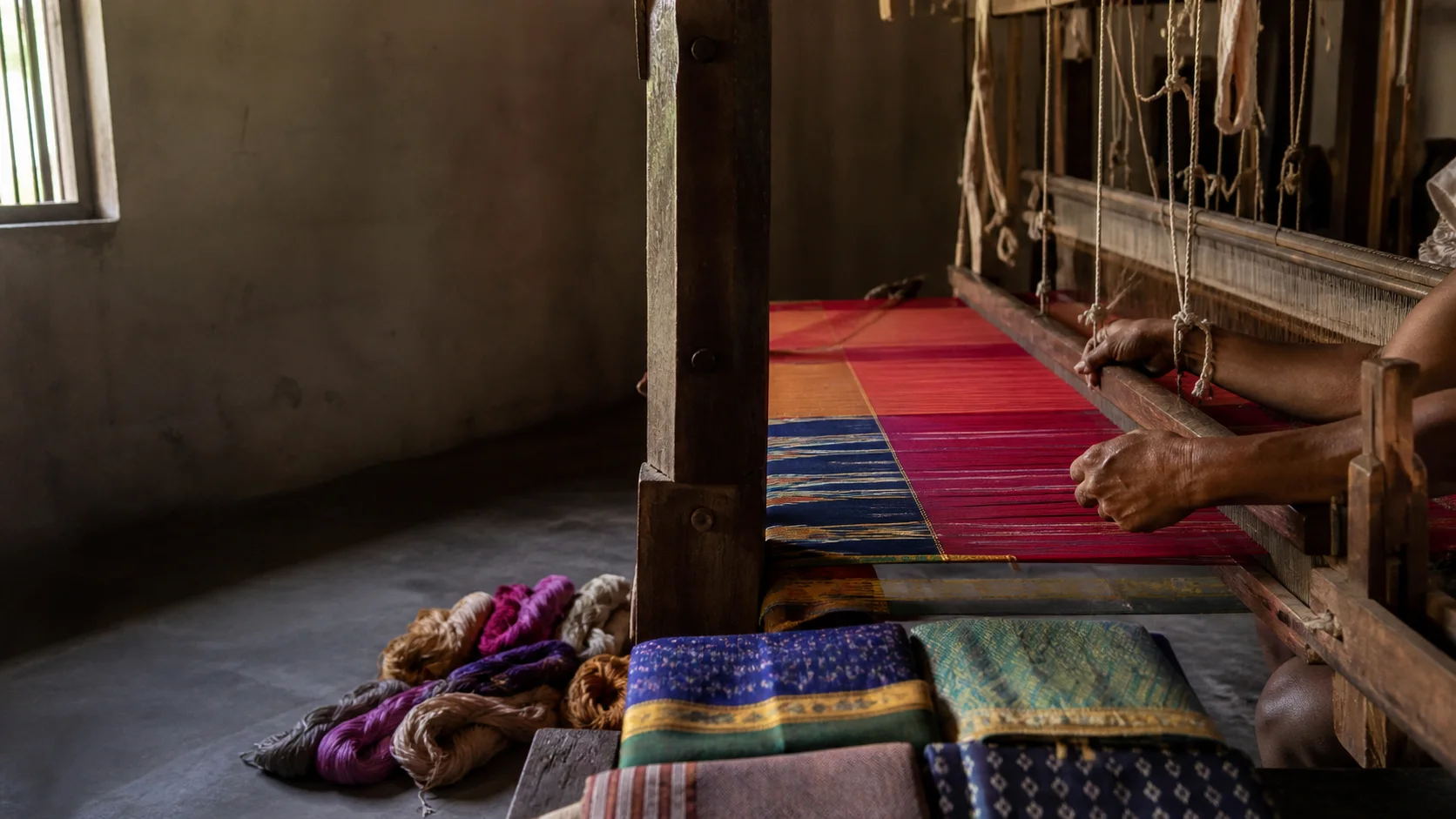 Realistic editorial illustration of a weaver working at a wooden loom with colourful textiles nearby